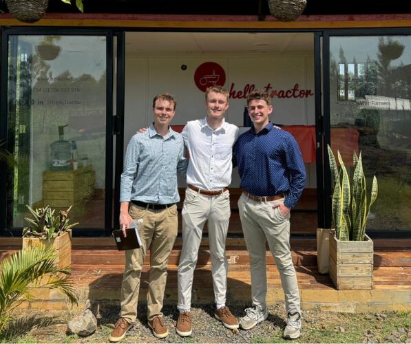 Three individuals pose for photo in front of building with sign that reads Hello Tractor