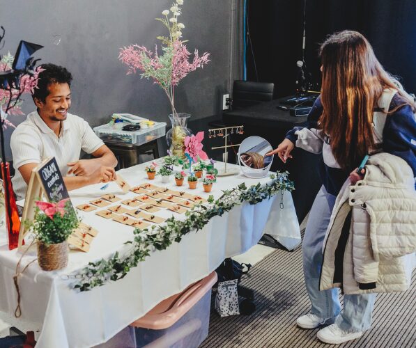 An individual sits at a table with items for sale on it, while another individual stands, looking at the items for sale