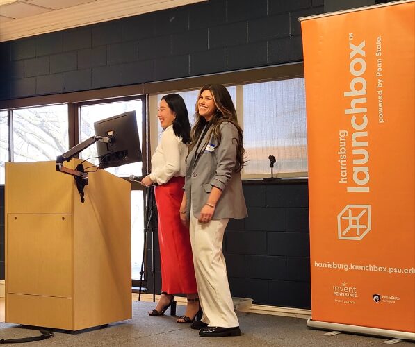 Two individuals stand on a stage with a Harrisburg LaunchBox standup banner behind them